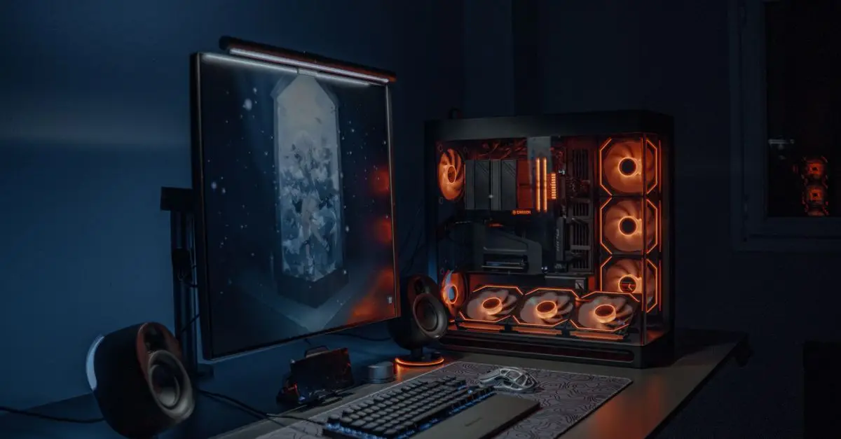 A gaming PC with orange fan lighting next to a monitor in a dark room